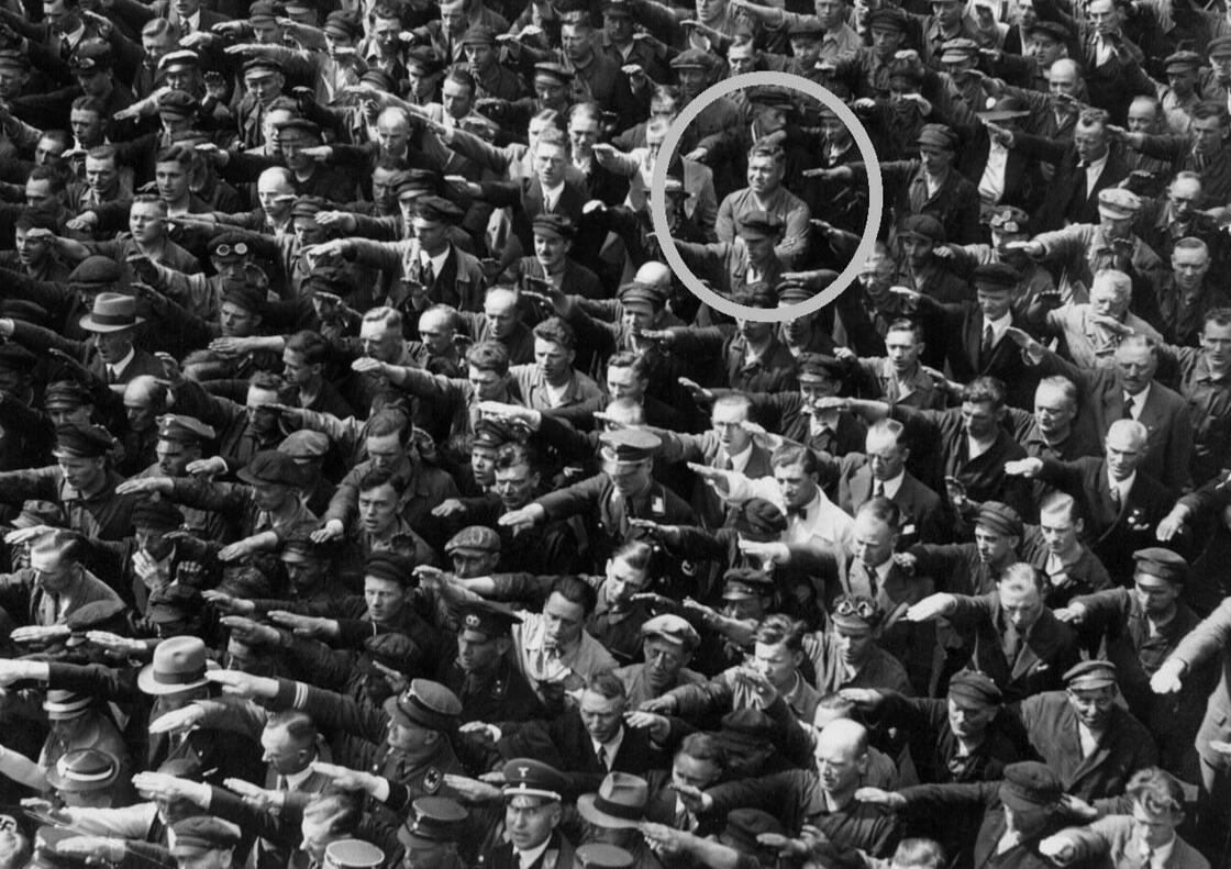 ภาพขาวดำปี 1936 ฝูงชนที่อู่ต่อเรือในฮัมบูร์กกำลังยกมือทำความเคารพแบบนาซี มีชายคนหนึ่งในวงกลม (เชื่อกันว่าคือ August Landmesser) ยืนกอดอกไม่ยอมยกมือ